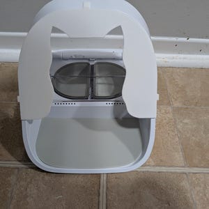 May include: A white, enclosed cat litter box with a rounded design. The top features a cat-ear-shaped flap that opens to reveal two clear, circular compartments. The base has a pull-out tray for easy cleaning.