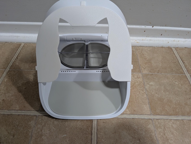 May include: A white, enclosed cat litter box with a rounded design. The top features a cat-ear-shaped flap that opens to reveal two clear, circular compartments. The base has a pull-out tray for easy cleaning.