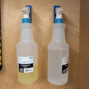 Puede incluir: Dos botellas de spray de plástico transparente con tapas azules y boquillas de pulverización blancas están montadas en una pared con soportes de plástico azules. Las botellas están etiquetadas como "Ironing Spray Bottle" y tienen boquillas de pulverización ajustables.