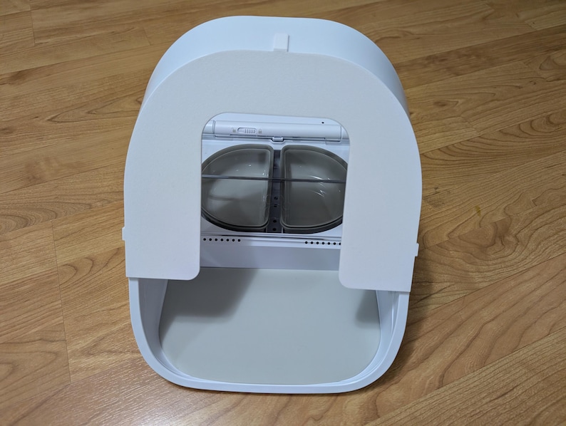 May include: A white, modern pet food dispenser with a curved top and a lower tray. The dispenser has two food compartments visible through a clear window. The unit is sitting on a wooden floor.