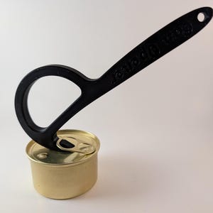 Puede incluir: Un abrelatas negro con las palabras "Can Opener" impresas en el mango. El abrelatas está colocado sobre una pequeña lata dorada. El abrelatas tiene un lazo y un mango con un agujero en el extremo.