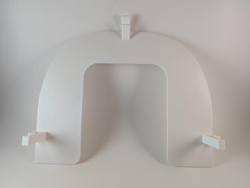 May include: A white, U-shaped, decorative shelf with a rectangular opening in the center. The shelf has a flat top and bottom with two small rectangular supports extending from the sides. A small rectangular piece extends from the top.