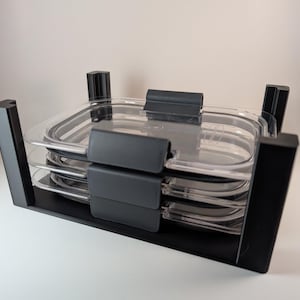 Puede incluir: Un juego de recipientes apilables para guardar alimentos. El juego incluye recipientes transparentes de forma ovalada con tapas y asas grises, sujetos dentro de un marco negro rectangular. Los recipientes parecen ser de plástico.