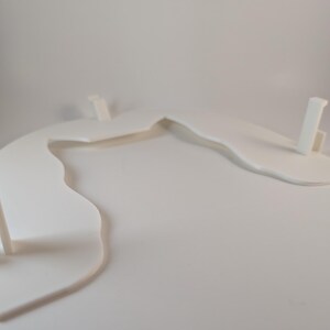May include: A white, abstract, curved display stand with three vertical supports. The stand has a flowing, organic shape, and is made of a solid, opaque material. The design is minimalist and modern, suitable for showcasing items.