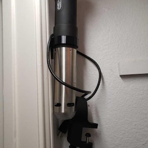 May include: A black and silver immersion circulator with a clamp attached to a wall. The circulator has a digital display and a cord.