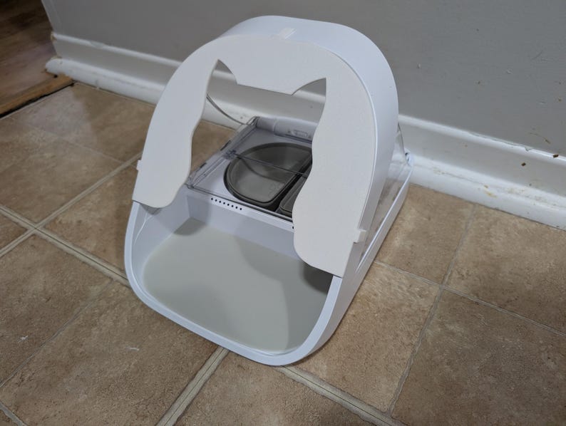 May include: A white pet food dispenser with a curved top and a clear food tray. The dispenser has a large, open base and a clear, removable food tray. The dispenser is designed to hold and dispense pet food.