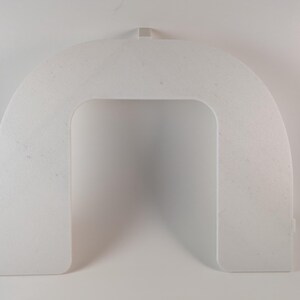 May include: A white, arched, plastic object with a rectangular opening in the center. The object has a small tab at the top and two small rectangular protrusions at the bottom. The surface has a slightly textured appearance.