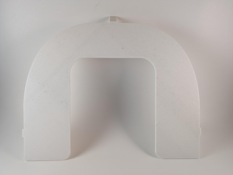 May include: A white, arched, plastic object with a rectangular opening in the center. The object has a small tab at the top and two small rectangular protrusions at the bottom. The surface has a slightly textured appearance.
