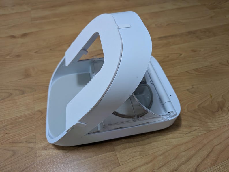May include: A white, modern pet food dispenser with a curved handle. The dispenser is open, revealing a clear container and internal components. The design is sleek and minimalist, suggesting a focus on functionality and ease of use. The dispenser is on a wooden surface.