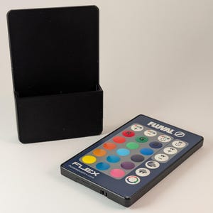 Puede incluir: Un soporte de pared de plástico negro para un mando a distancia. El mando a distancia es azul y tiene el logotipo de Fluval y botones para controlar las luces del acuario. Los botones están etiquetados con colores, incluidos rojo, verde, azul, blanco y un icono de luna.