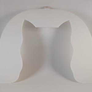 May include: A white, arched, foam-like object with a rectangular top and curved sides. The object has a cutout in the center and a small rectangular tab at the top. The sides have a wavy, organic shape.