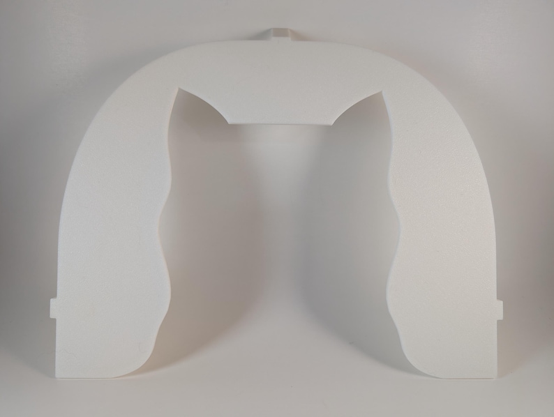 May include: A white, arched, foam-like object with a rectangular top and curved sides. The object has a cutout in the center and a small rectangular tab at the top. The sides have a wavy, organic shape.