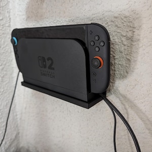 May include: A black wall-mounted dock holding a Nintendo Switch console. The console features black Joy-Con controllers with one orange joystick and one blue joystick. The dock displays the Nintendo Switch logo and is connected to black power cables.