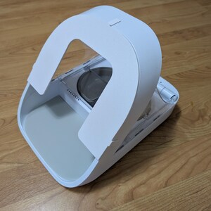 May include: A white pet food dispenser with a curved handle and a gray interior. The dispenser is on a wooden floor. The design is modern and minimalist.