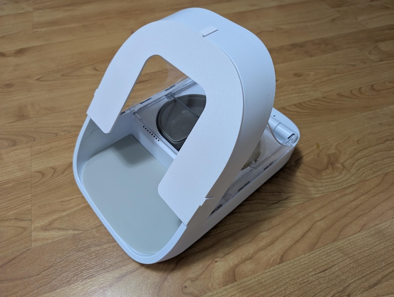 May include: A white pet food dispenser with a curved handle and a gray interior. The dispenser is on a wooden floor. The design is modern and minimalist.