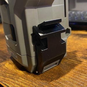 Può includere: Un primo piano di un fermo in plastica nera su una custodia in plastica grigia. Il fermo è fissato alla custodia con una piccola vite.