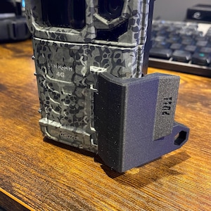 May include: A camouflage-patterned trail camera with a black mounting bracket. The camera has a black lens and the text "LINK-WM 4G" and "SPOT POINT" printed on the side.