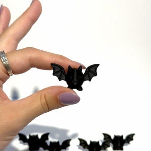 May include: A small, matte black figurine shaped like a bat with pointed ears and outstretched wings. The bat has a rounded body and a smooth, solid surface texture. Several identical black bat figurines are arranged on a plain white surface.