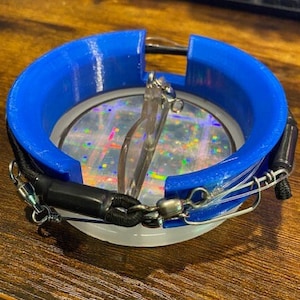 Op de afbeelding: Een blauw en wit cirkelvormig apparaat met een helder, iriserend midden. Het apparaat heeft een blauwe, 3D-geprinte buitenring en een zwart koord met metalen sluitingen. Een heldere bril zit in het apparaat.