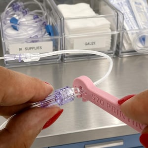 Peut inclure: Un outil clé en plastique rose clair avec le texte 570 PRINTING en relief sur le côté, utilisé pour serrer un connecteur IV en plastique transparent. L'outil présente une ouverture en forme de U conçue pour s'adapter autour du tube IV.