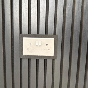 May include: A double electrical socket with two switches, set in a dark grey frame. The socket is mounted on a vertical, grey-toned wooden panel wall.