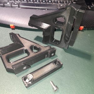 May include: Black 3D-printed components, possibly for a mechanical assembly, are arranged on a green surface. The parts include angled brackets with rectangular and circular openings, a small rectangular piece, and a screw. The components are near a laptop keyboard.