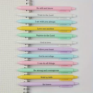 May include: A selection of pastel-coloured pens with inspirational quotes printed on them. The pens are arranged vertically on a spiral notebook. Each pen has a different colour and a different message, such as "Be still and know" and "Trust in the Lord."