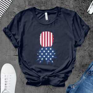 May include: A navy blue t-shirt with a pineapple design featuring the American flag colors. The pineapple is made up of red and white stripes with a blue star pattern on the bottom.
