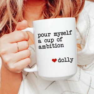 Puede incluir: Una taza de cerámica blanca con el texto impreso en negro "pour myself a cup of ambition" seguido de un pequeño icono de corazón rojo y la palabra "dolly" en negro. La taza tiene un asa estándar y un diseño cilíndrico liso.