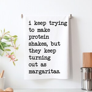 May include: White kitchen towel with the text "i keep trying to make protein shakes, but they keep turning out as margaritas" in black font. The towel is hanging on a silver bar.