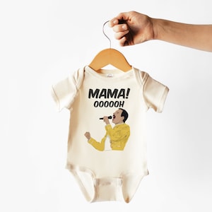 May include: A cream-colored baby onesie with a black and yellow illustration of Freddie Mercury singing with the text "MAMA! OOOOOOOH".