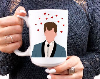 May include: White ceramic mug featuring a stylized illustration of a person in a blue suit with a bolo tie, surrounded by red hearts. The mug has a large handle and is held by a person with white nail polish.