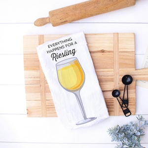 May include: White kitchen towel with the text "Everything Happens for a Riesling" and a graphic of a wine glass filled with yellow liquid. The towel is on a wooden cutting board with a rolling pin and measuring spoons.