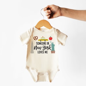 Könnte beinhalten: Ein cremefarbener Baby-Strampler mit einer schwarzen Grafik eines gelben Taxis, des Empire State Building, der Freiheitsstatue, eines Brezels und eines Apfels. Der Text auf dem Strampler lautet "Someone in New York loves me."