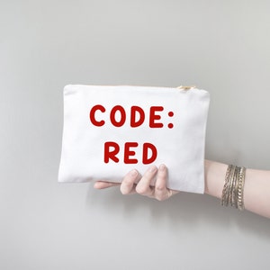 以下が含まれることがあります： 手で持たれた、金色のジッパーが付いた白いキャンバス地のポーチ。ポーチには赤い文字で「CODE: RED」と書かれています。長方形のポーチは柔らかい生地でできています。背景は無地のライトグレーです。