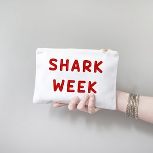 Op de afbeelding: Witte canvas pouch met een gouden rits, met de rode tekst "SHARK WEEK". De pouch wordt vastgehouden door een hand, met zilveren armbanden om de pols. De achtergrond is effen lichtgrijs.