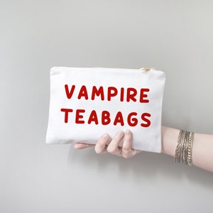 May include: A white canvas pouch with a gold zip, held by a hand. The pouch features the words "VAMPIRE TEABAGS" in bold red letters. The person is wearing multiple silver bangles.