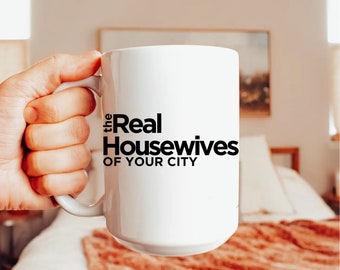 May include: White ceramic mug with the text "The Real Housewives of YOUR CITY" in black. The mug is held by a hand, with a blurred background of a bedroom setting.