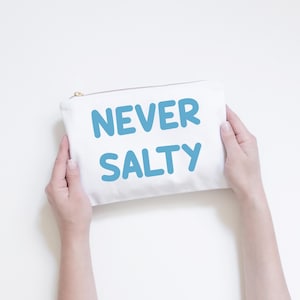 May include: A white canvas pouch with a gold zip, held by two hands. The pouch has the text "NEVER SALTY" in large, light blue letters. The background is plain white.
