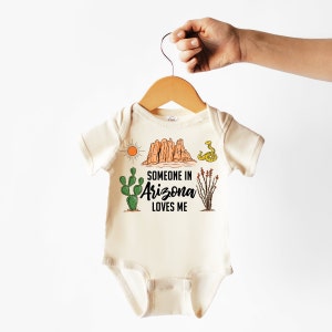 May include: A cream-colored baby onesie with a graphic design featuring a desert landscape, cactus, snake, and the text "Someone in Arizona loves me."