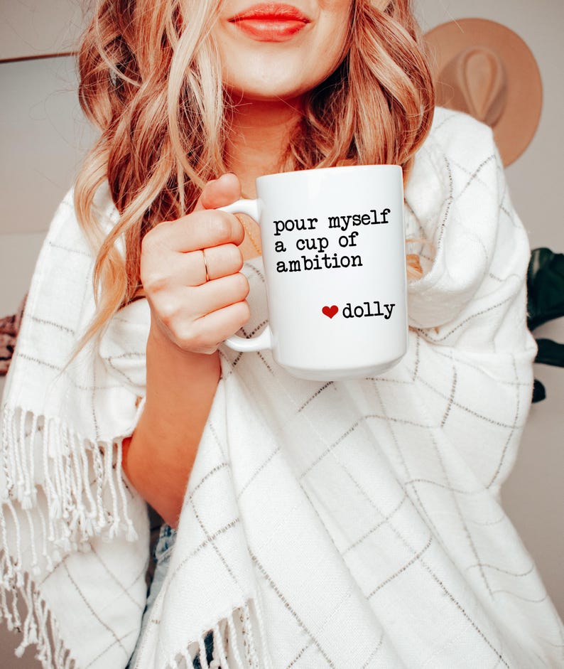 Może przedstawiać: Biały ceramiczny kubek z czarnym nadrukiem "pour myself a cup of ambition", po kt&oacute;rym następuje mała czerwona ikona serca i słowo "dolly" w kolorze czarnym. Kubek ma standardowy uchwyt i gładki, cylindryczny kształt.