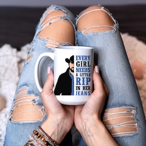 May include: White ceramic mug featuring a graphic of a man in a cowboy hat and the text "EVERY GIRL NEEDS A LITTLE RIP IN HER JEANS". The mug is held by a person wearing ripped blue jeans.