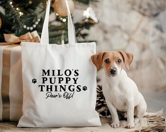 May include: A white canvas tote bag featuring the black printed text "MILO'S PUPPY THINGS Paw's Off!" with two small paw print icons. The bag has two long shoulder straps and is positioned next to a small white and brown puppy.
