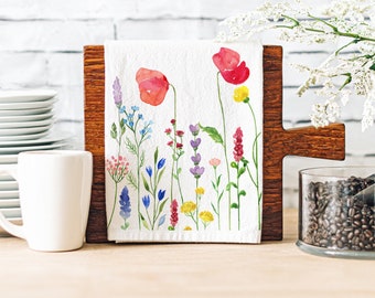 May include: A white tea towel with a watercolour floral design. It features red poppies, blue, purple, and yellow wildflowers. The towel is displayed on a wooden chopping board. A white mug and plates are in the background.