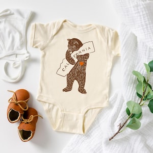 Puede incluir: Un body de bebé de color crema con un oso marrón abrazando un contorno marrón del estado de California con el texto "CALIFORNIA" en él. El oso tiene un corazón rojo en el pecho.
