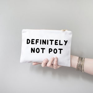 Könnte beinhalten: Eine weiße Canvas-Tasche mit goldenem Reißverschluss, vor grauem Hintergrund gehalten. Auf der Tasche steht in fetten, schwarzen Buchstaben "DEFINITELY NOT POT". Das Bild zeigt auch ein Handgelenk mit mehreren silbernen Armbändern.