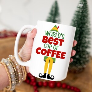 May include: White ceramic coffee mug with the text "WORLD'S BEST CUP OF COFFEE" in red and green. The mug features an elf design with yellow legs, green tunic, and black shoes. A festive holiday-themed mug.
