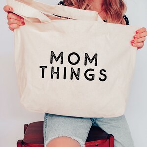 Puede incluir: Bolso tote de lona color crema con las palabras "MOM THINGS" impresas en letras negras en negrita. El bolso tiene asas color crema y es sostenido por una persona.