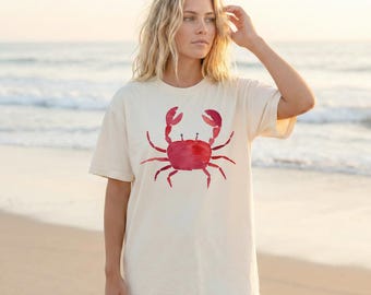 Puede incluir: Camiseta color crema con un diseño de cangrejo rojo estilo acuarela. El cangrejo tiene un cuerpo y pinzas detallados. La camiseta es de corte holgado, perfecta para uso casual. El fondo muestra una escena de playa con el océano y arena.