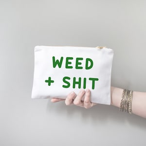 May include: White canvas pouch with a gold zipper, held by a hand. The pouch features the words "WEED + SHIT" in bold green letters. The person holding the pouch is wearing multiple silver bracelets.
